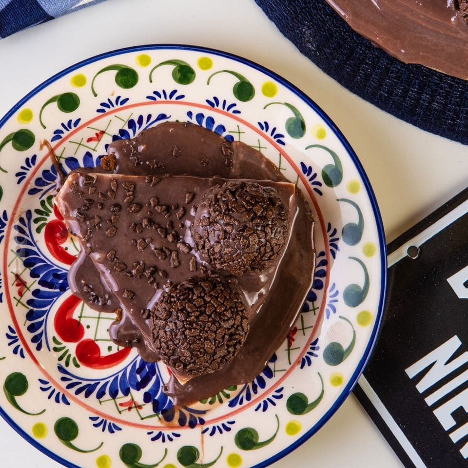 Apresentamos nossa suntuosa Fatia de Torta de Fior di Latte com Brigadeiro. Um prato que combina a cremosidade do autêntico sorvete italiano Fior di Latte com a doçura inconfundível de um clássico brigadeiro ao leite brasileiro. A cobertura é generosamente polvilhada com granulado de chocolate belga, conhecido mundialmente pela sua qualidade superior. A base, feita de biscoito amantegado, proporciona a textura crocante perfeita que contrasta maravilhosamente com a suavidade do sorvete. Uma sinfonia de sabores e texturas que promete encantar seu paladar. Experimente e se apaixone por essa delícia gelada e irresistível.