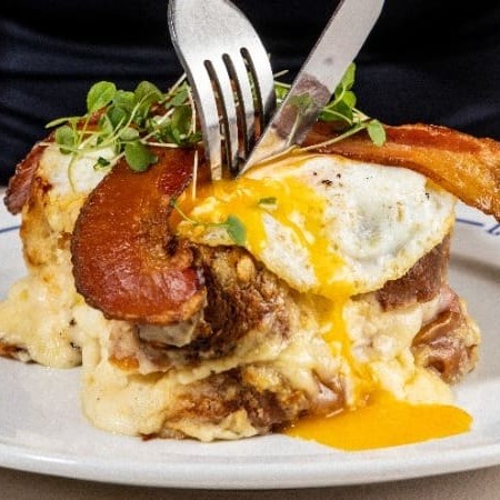 Sanduiche no pão brioche, queijo, presunto Royale, molho bechamel, gratinado no forno, com um ovo frito e fatias de bacon defumando crocante.