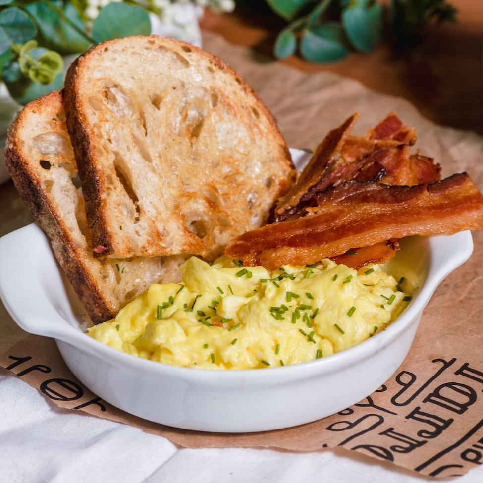 Ovos mexidos cremosos com fatias de bacon. Acompanham uma fatia do nosso pão Sourdough.