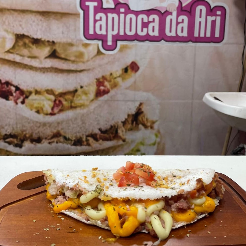 Uma Das Mais Pedidas! Diretamente da Feira Nacional do Podrão para o seu prato, a Tapi Tudo da Ari é um verdadeiro espetáculo de sabor!   Uma Mega tapioca recheada com calabresa suculenta, presunto saboroso, cheddar cremoso, requeijão derretido, mussarela puxa-puxa, bacon, ovos de codorna, cebola, tomate fresquinho, orégano perfumado e alho torrado crocante!  Se você gosta de uma tapioca bem recheada e cheia de sabor, essa é pra você! Peça agora e descubra por que ela fez tanto sucesso! ️