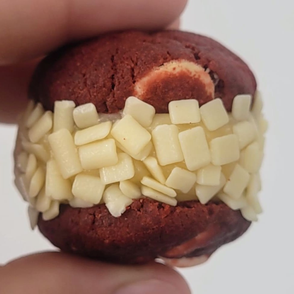 Sanduichinho com 2 mini cookies red velvets feitos com cacau 100% e chocolate branco, recheado com brigadeiro cremoso e finalizado no confeito branco Uma explosão de sabores