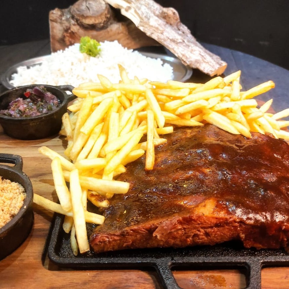Desfrute da nossa suculenta costela suína de aproximadamente 550g, cuidadosamente preparada com o nosso dry rub especial e defumada por 6 horas na lenha frutífera. Ela é servida com farofa de bacon crocante, molho chimichurri, 500g de arroz e a sua escolha de 500g de batata ou aipim. Escolha abaixo os seus acompanhamentos