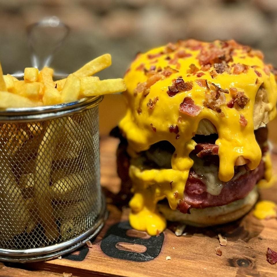 200 gramas de hamburguer de carne suína recheado com queijo mussarela, cebola caramelizada no vinho, cheddar, bacon e pão com cobertura de cheddar e crispy de bacon. Acompanha: Fritas.  (A cobertura de cheddar + bacon para delivery é enviado em pote separado)