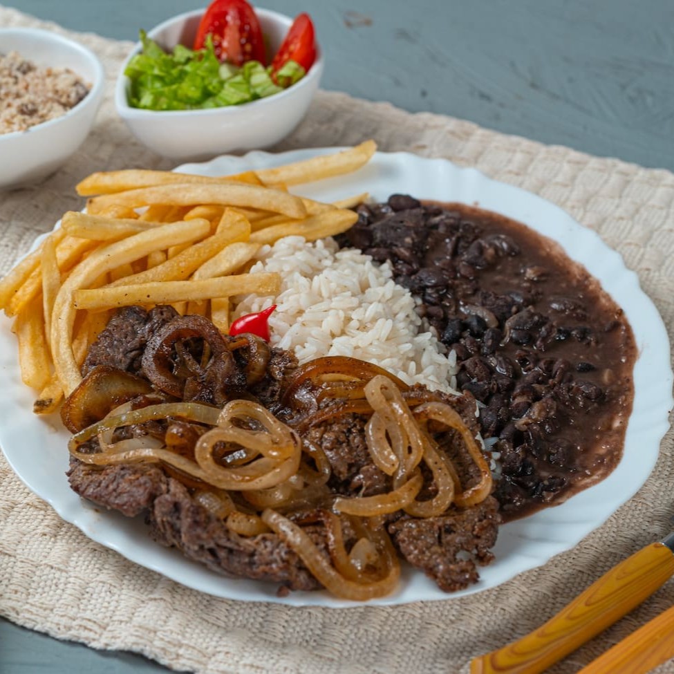 Suculento bife grelhado no ponto perfeito, coberto com cebolas douradas e bem temperadas. Acompanha arroz soltinho, feijão caseiro saboroso, batatas fritas crocantes, farofa temperada e uma salada fresquinha para equilibrar o prato. Uma refeição clássica e cheia de sabor!