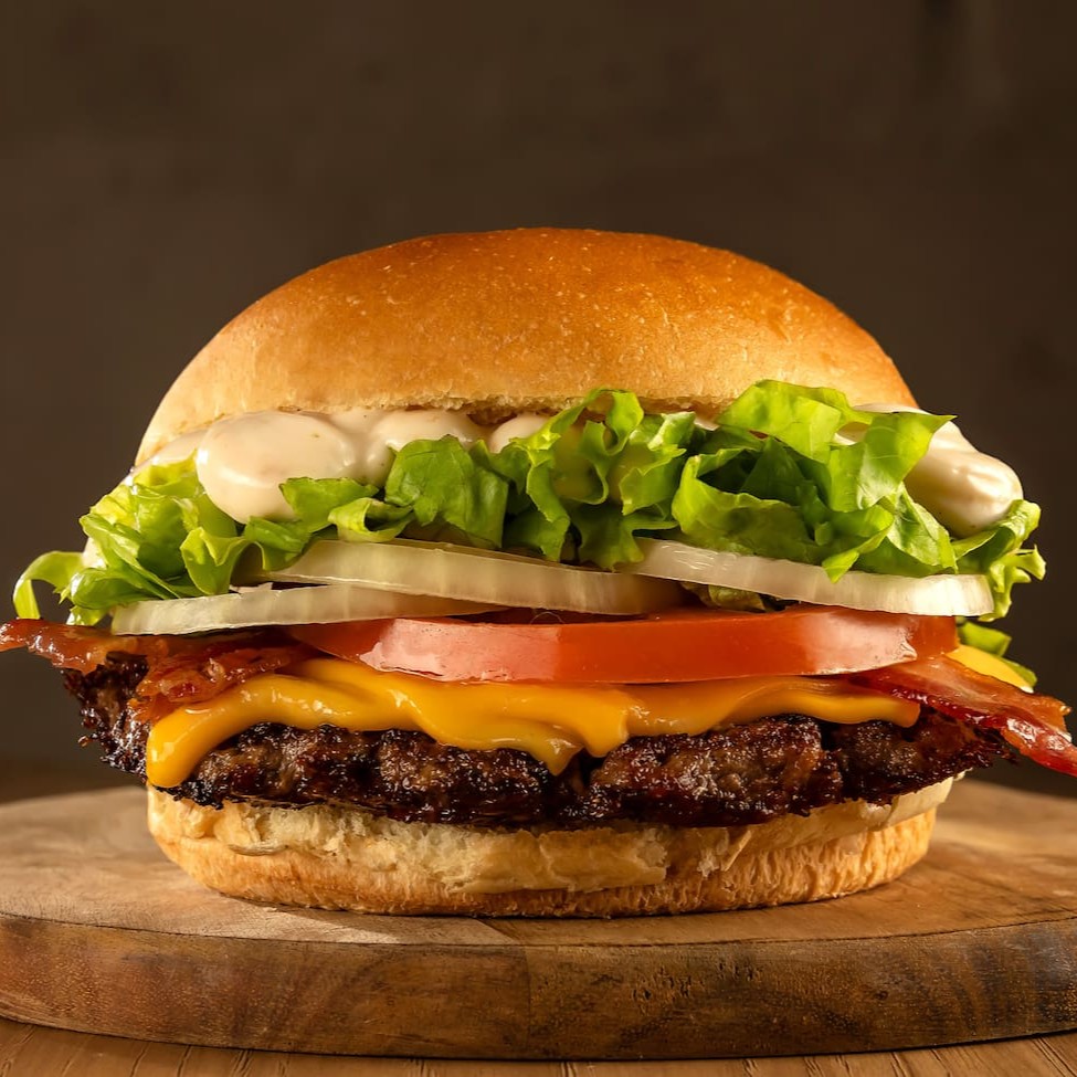 Delicioso Burger de carne black angus de 80g prensado na chapa, coberto com queijo prato, bacon crocante, alface, tomate, cebola e molho da casa à base de alho no pão brioche.