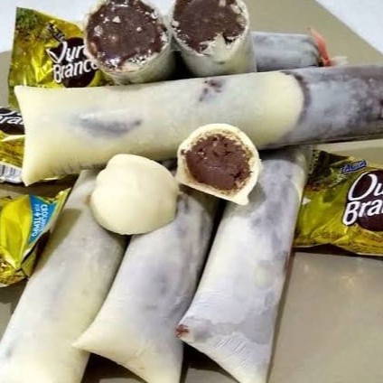 O sacolé de Ouro Branco é uma explosão de sabores e texturas, inspirado no famoso bombom. A base é um cremoso sorvete de chocolate, intenso e aveludado, que derrete na boca, trazendo aquele gostinho irresistível de cacau. Misturados ao sorvete, pedaços crocantes de castanha adicionam um toque especial, equilibrando o doce com uma leve crocância. Para finalizar, uma casquinha de chocolate branco envolve o sacolé, formando uma camada fina e quebradiça que se parte delicadamente a cada mordida, liberando todo o sabor do recheio. Uma combinação perfeita entre cremosidade, crocância e o sabor marcante do chocolate!