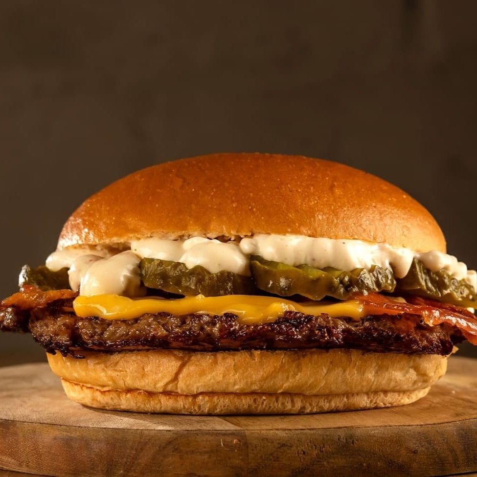 Delicioso Burger de carne black angus de 80g prensado na chapa, coberto com queijo prato, picles, bacon crocante e molho da casa à base de alho no pão brioche.