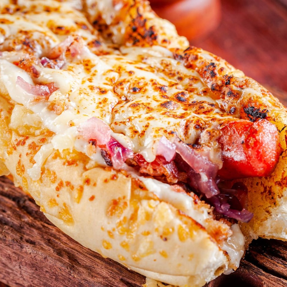 Linguiça artesanal de pernil com alho,  creme de queijos , cebola caramelizada, farofa de bacon,  muçarela maçaricada,  na baguete de parmesão.