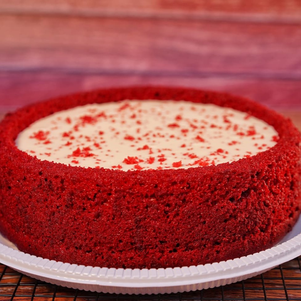 Apresentamos o nosso 'Red Velvet com Leite Ninho', uma verdadeira obra-prima da nossa categoria 'Bolo Piscina'. Este bolo, de 1,2 kg e capaz de servir até 16 fatias generosas, é uma combinação harmoniosa de uma massa branca finamente elaborada, delicadamente tingida com corante alimentício de alta qualidade para obter a icônica cor vermelha. A cobertura, uma camada indulgente de Leite Ninho, adici