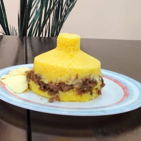 Cuscuz de milho recheado com carne desfiada, manteiga de leite e muçarela. Parte da Manteiga vai incorporada ao recheio e parte vai separadamente em um potinho.