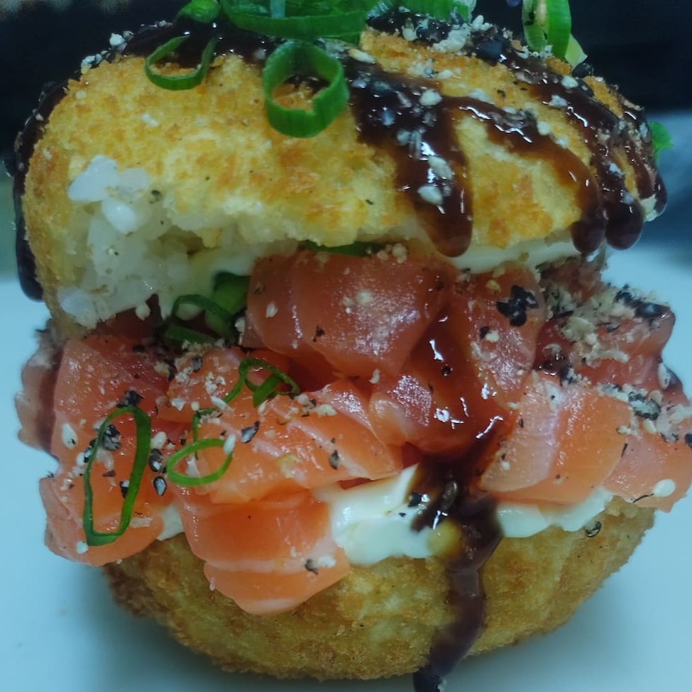 Hambúrguer feito base de arroz empanado e frito, cream cheese caprichado, salmão fresco em cubinhos, gergelim torrado e moído, cebolinha e um pouco de teriaki por cima