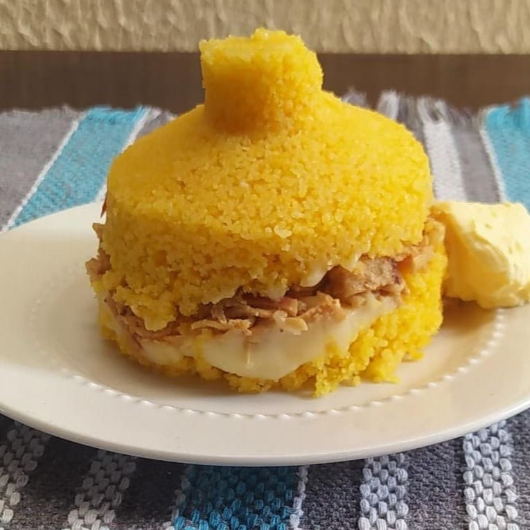 Cuscuz de milho recheado com frango desfiado, manteiga de leite e requeijão. Parte da Manteiga vai incorporada ao recheio e parte vai separadamente em um potinho.
