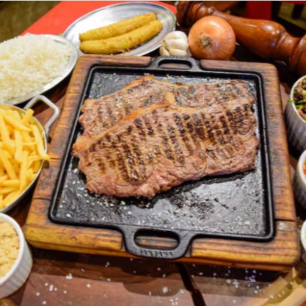 Desfrute da nossa '111 - Alcatra com Guarnição - 2 Pessoas', um requintado prato da categoria 'Churrasco na Pedra'. A alcatra, suculenta e bem temperada, é acompanhada por um arroz soltinho e um feijão tropeiro tradicional, repleto de sabor. As batatas fritas são crocantes e douradas, trazendo um toque de textura ao prato. O molho suave, a farofa saborosa e a banana frita, doce e macia, complementam a experiência gastronômica. Para finalizar, um creme de alho especial com ervas finas, perfumado e cremoso, adiciona um toque sofisticado. Um prato completo para compartilhar em boa companhia!