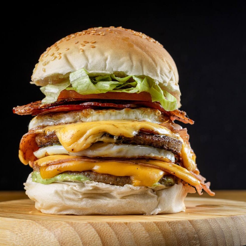 X-tudo duplo, pra quem tem muita fome: pão macio, 2 carnes suculentas, 2 queijos, 2 presuntos, bacon crocante, 2 ovos, salada e molho verde. Pesado, completo e cheio de axé — o maior da categoria, sem miséria!