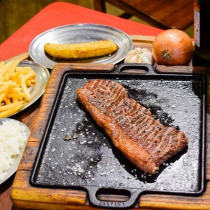 Desfrute de uma suculenta alcatra, perfeitamente grelhada na pedra, acompanhada por uma saborosa guarnição destinada a encantar o seu paladar. A combinação inclui um arroz branco leve e soltinho, um autêntico feijão tropeiro, batatas fritas crocantes e um molho temperado criteriosamente. A farofa, rica e saborosa, e a banana frita, adocicada e macia, adicionam um contraste delicioso ao prato. Finalizando com um toque especial, um creme de alho requintado, preparado com ervas finas, aprimorando a experiência culinária. Este prato é perfeito para satisfazer o apetite de uma pessoa, oferecendo uma refeição completa e indulgente.