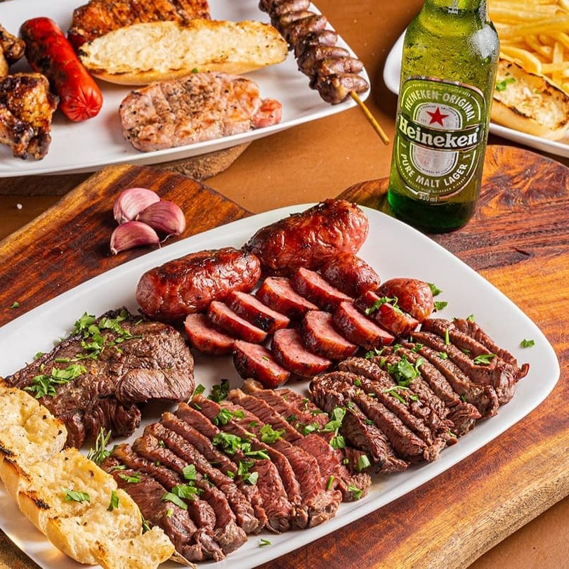 "4 linguiças, 4 drumets, 10 unidades de coração, em média 200g de suíno, 1 salsichão, em média 450g de coração da alcatra e 300g de batata frita. *Acompanhamento= Pão de alho, molho vinagrete e farofa*   ""Porção generosa e irresistível que reúne 4 linguiças suculentas, 4 drumets douradinhos, 10 unidades de coração saboroso, cerca de 200g de carne suína macia, 1 salsichão especial e aproximadamente 450g de coração de alcatra bem temperado. Para acompanhar essa combinação repleta de sabor, servimos também 300g de batata frita crocante, além do pão de alho douradinho, molho vinagrete fresquinho e farofa crocante e temperada. Uma experiência completa para quem quer se deliciar com o melhor da carne e acompanhamentos tradicionais, perfeita para compartilhar ou saborear com vontade!"""