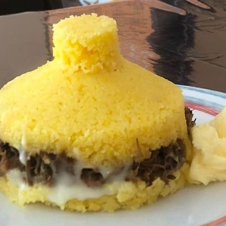 Cuscuz de milho recheado com carne desfiada, manteiga de leite e requeijão. Parte da Manteiga vai incorporada ao recheio e parte vai separadamente em um potinho.