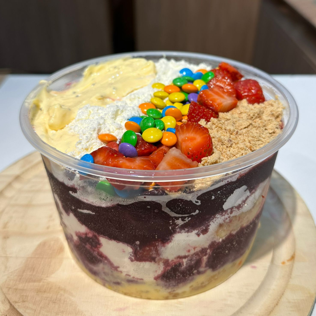 Açaí tradicional super cremoso com a leve acidez da mousse de maracujá, pedaços fresquinhos de morango, a crocância da paçoca, disqueti colorido e finalizado com leite em pó.