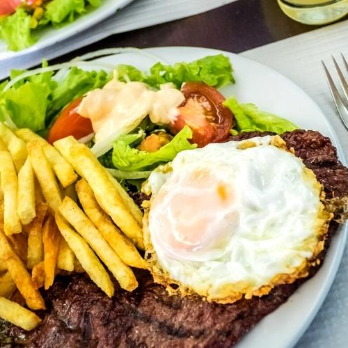 A refeição é composta por, arroz, feijão, 1 bife médio, um ovo frito, uma porção p de polenta ou batata frita, uma porção p de salada mista e um refrigerante de lata.