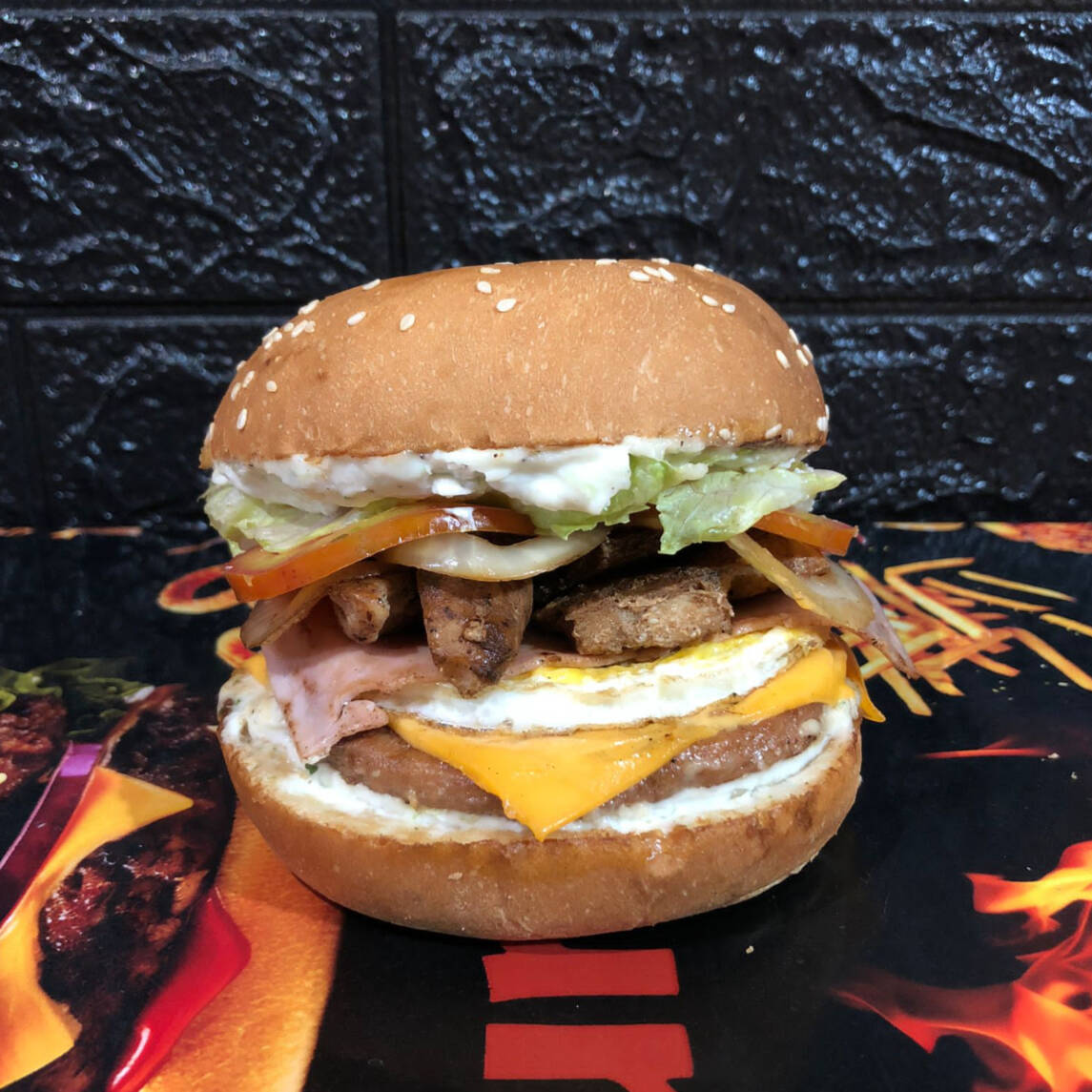 Pão com gergelim, carne industrial, queijo cheddar fatiado, ovo frito, presunto, alface americana, tomate, cebola e molho opcional.