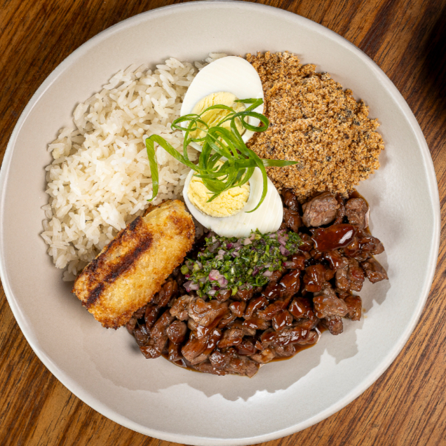 Filé mignon picado na ponta da faca com toque de demi-glace artesanal, feijão preto, arroz soltinho, farofa crocante de maracujá, banana empanada crocante assada ao forno e ovo.
