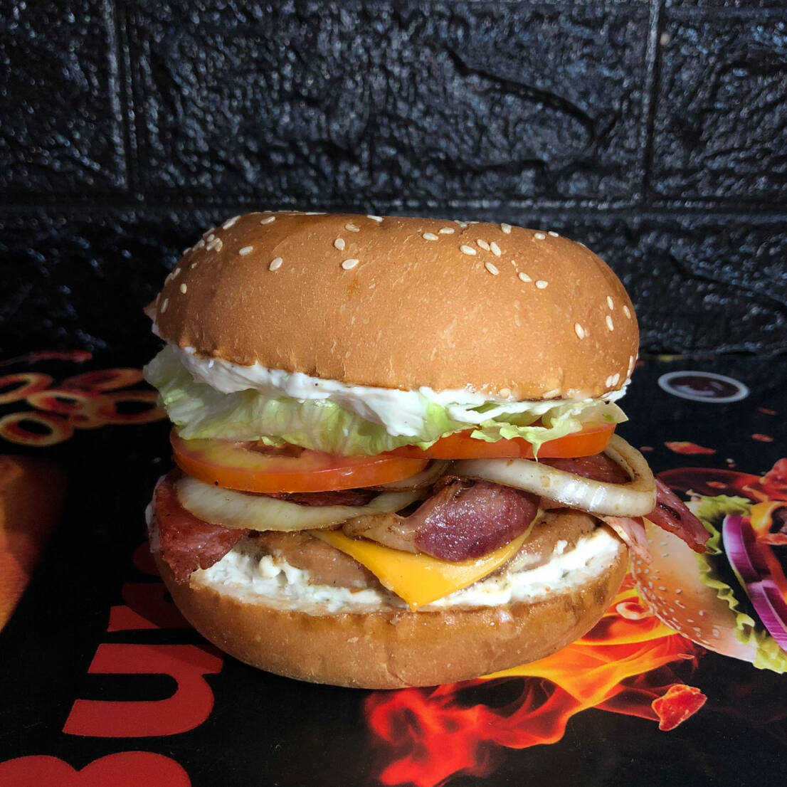 Pão com gergelim, carne industrial, bacon, queijo cheddar fatiado, presunto, alface americana, tomate, cebola e molho opcional.