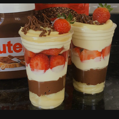 O Copo da Felicidade de Ninho com Morango e Nutella é uma sobremesa que une três sabores extremamente populares, criando uma experiência rica e equilibrada em cada colherada. A base é o creme de Leite Ninho, que traz uma textura aveludada e um sabor doce e reconfortante, remetendo à infância. Ele é intercalado com pedaços de morangos frescos, que oferecem um toque de acidez e umidade, quebrando o dulçor e adicionando uma leveza frutada. Para coroar a mistura, a Nutella entra com sua intensidade de chocolate e avelã. A ganache de Nutella adiciona um sabor marcante e uma textura mais densa, criando um contraste perfeito com a delicadeza dos outros dois elementos. O resultado é uma sobremesa visualmente atraente, com camadas de branco, vermelho e marrom, e um sabor complexo que transita entre o doce, o frutado e o rico. É uma combinação que agrada a todos e se tornou um clássico da confeitaria brasileira.