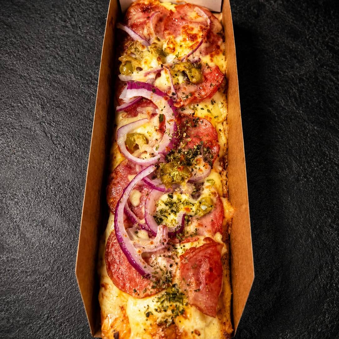 Formato: Retangular, sugerindo que foi assada em uma assadeira ou é um estilo conhecido como pizza à metro ou pizzeta. Base de Queijo: Há uma generosa camada de queijo muçarela (ou similar) derretida por baixo dos outros ingredientes, com as bordas tostadas, indicando que foi bem assada. calabresa: fatia de calabresa cozido e que parecem ter sido temperados com ervas secas antes de serem colocados na pizza. Cebola: Fatias finas e cruas de cebola branca estão espalhadas sobre o frango e o queijo, adicionando um toque de frescor e sabor. Tempero: É visível uma quantidade de ervas secas (como orégano ou mix de ervas finas) polvilhada sobre a cobertura, contribuindo para o aroma e sabor.