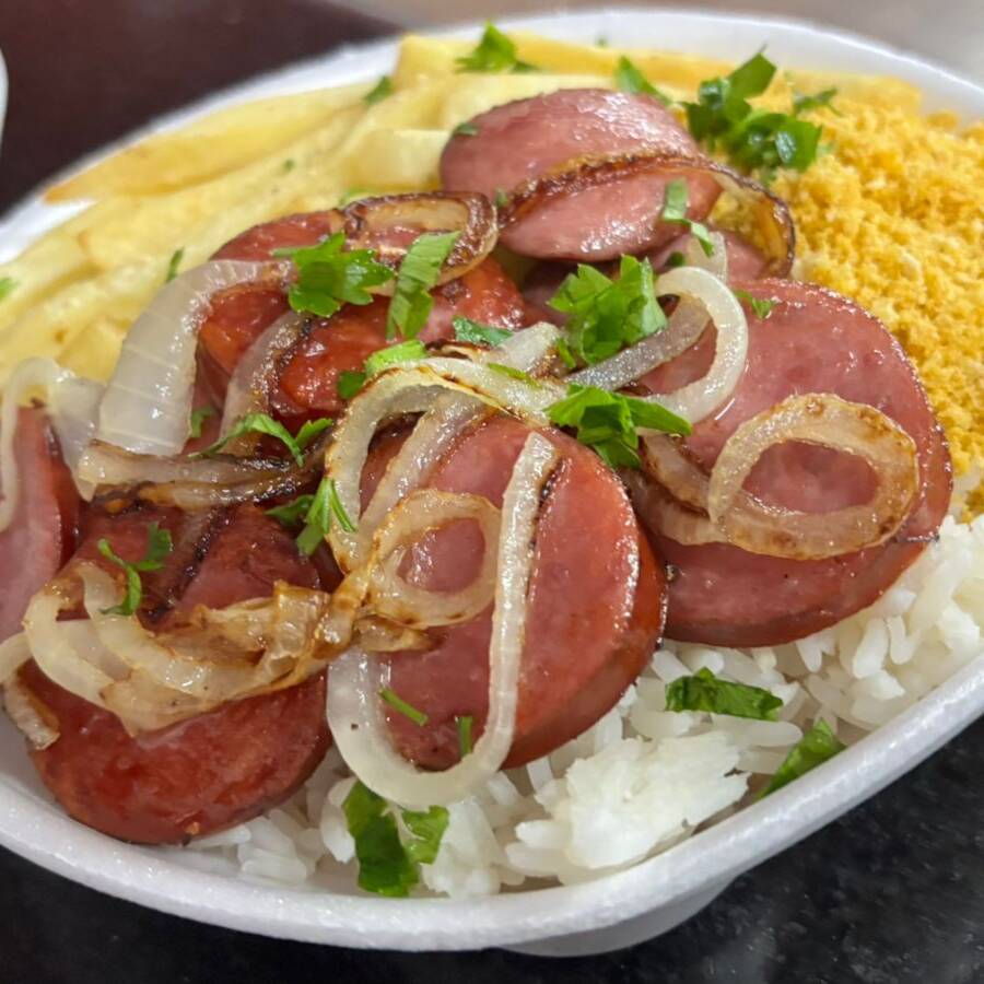 O prato de Calabresa Acebolada oferece fatias de linguiça calabresa douradas na frigideira, acompanhadas de cebolas caramelizadas, realçando o sabor defumado da linguiça. Junto a isso, o arroz branco soltinho e o feijão bem temperado trazem conforto e uma farofa crocante.
