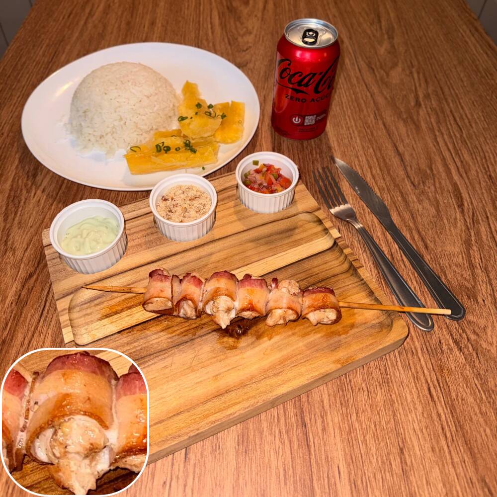 Espeto de medalhão de frango e bacon acompanhado por arroz, mandioca, vinagrete, farofa crocante e molho verde.