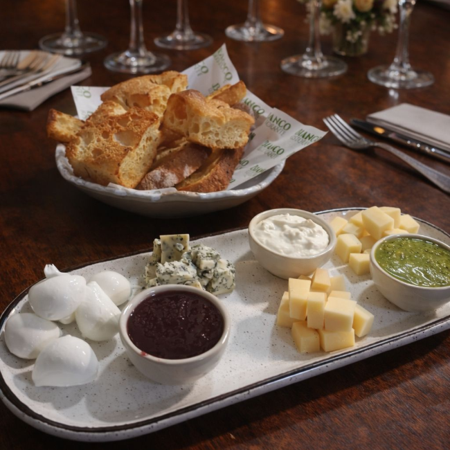 Seleção de queijos da casa: grana padano, gorgonzola, queijo de búfala, parmesão e mousse de burrata. Servido com pães da casa, pesto de manjerição e geleia de frutas.