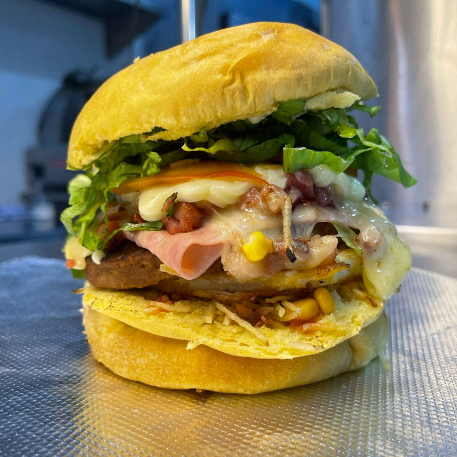 Pão prensado, hambúrguer, mussarela, presunto, ovo, bacon, ketchup, maionese, alface, tomate, catupiry, batata palha e milho.