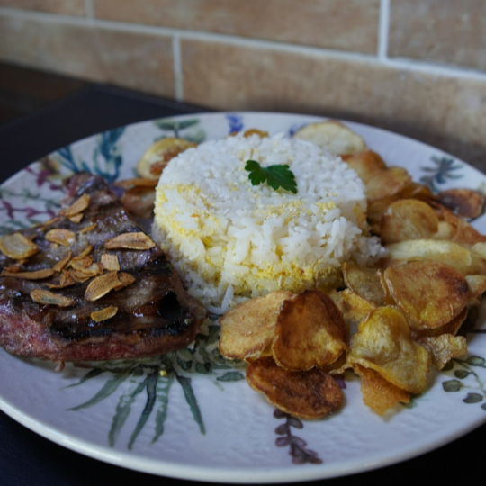 Bife de Vazio “Angus” com lâminas de alho, acompanhado de arroz com farofa crocante e batata chips.