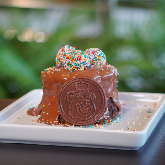 Bolo de chocolate 50%, recheado com brigadeiro com cobertura de chocolate ao leite derretido e enfeitado com a moeda do luguito e 3 brigadeiros coloridos.