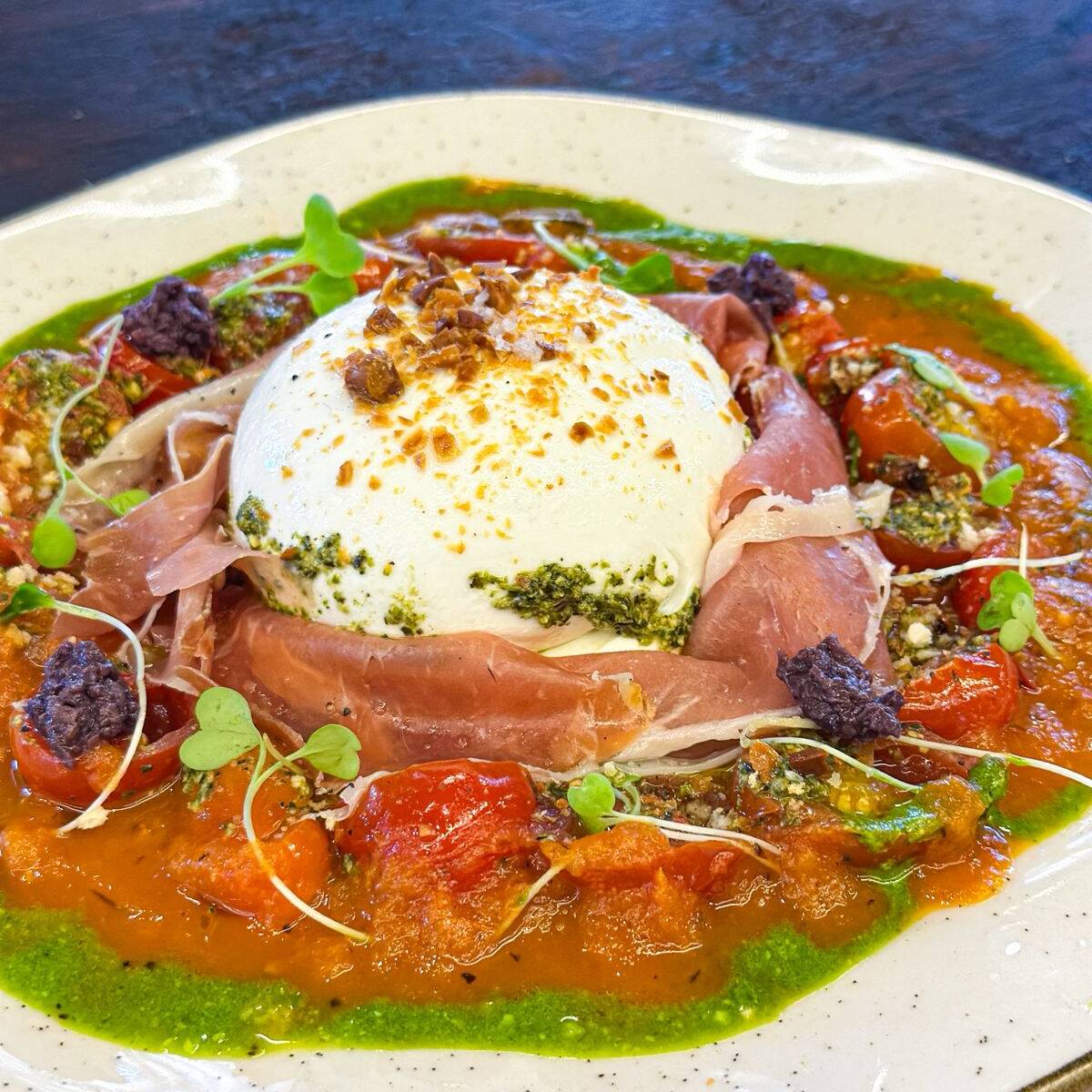 Burrata servida sob passata de tomate, tapenade de azeitonas pretas, presunto parma, castanha de baru, pesto de rúcula.