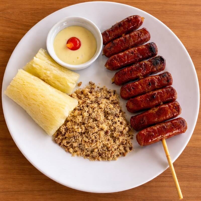 Espetinho de Linguiça de Costela Angus grelhado na brasa, farofa artesanal e mandioca cozida.