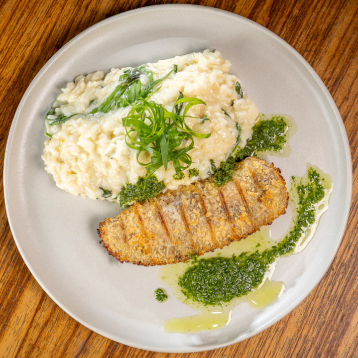 Tilápia empanada e assada ao forno com ervas, sobre risoto cremoso de limão siciliano e manjericão. Finalizado com redução cítrica de coentro e rúcula fresca.