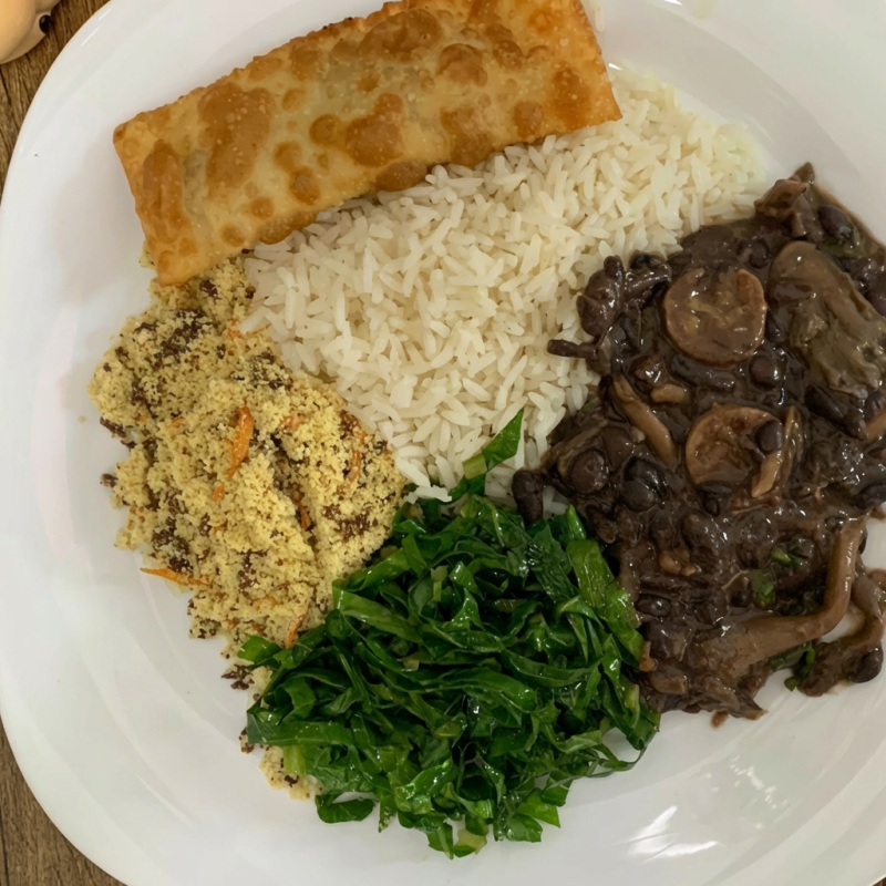 Feijoada vegana acompanha arroz branco, couve refogada, farofa de alho com proteína de bacon, molho de pimenta e pastelzinho recheado com pts. Feijoada recheada com shimeji e linguiça do futuro