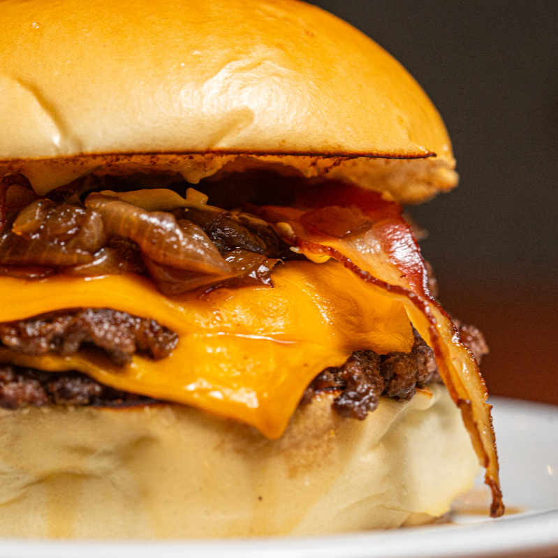 Dois blends bovinos de 100g prensados na chapa até formar aquela crosta dourada cheia de sabor. Montado com queijo cheddar derretido, bacon crocante, cebola caramelizada e a maionese da casa.
Um burger mais intenso, suculento e direto ao ponto, ideal pra quem quer comer bem de verdade.