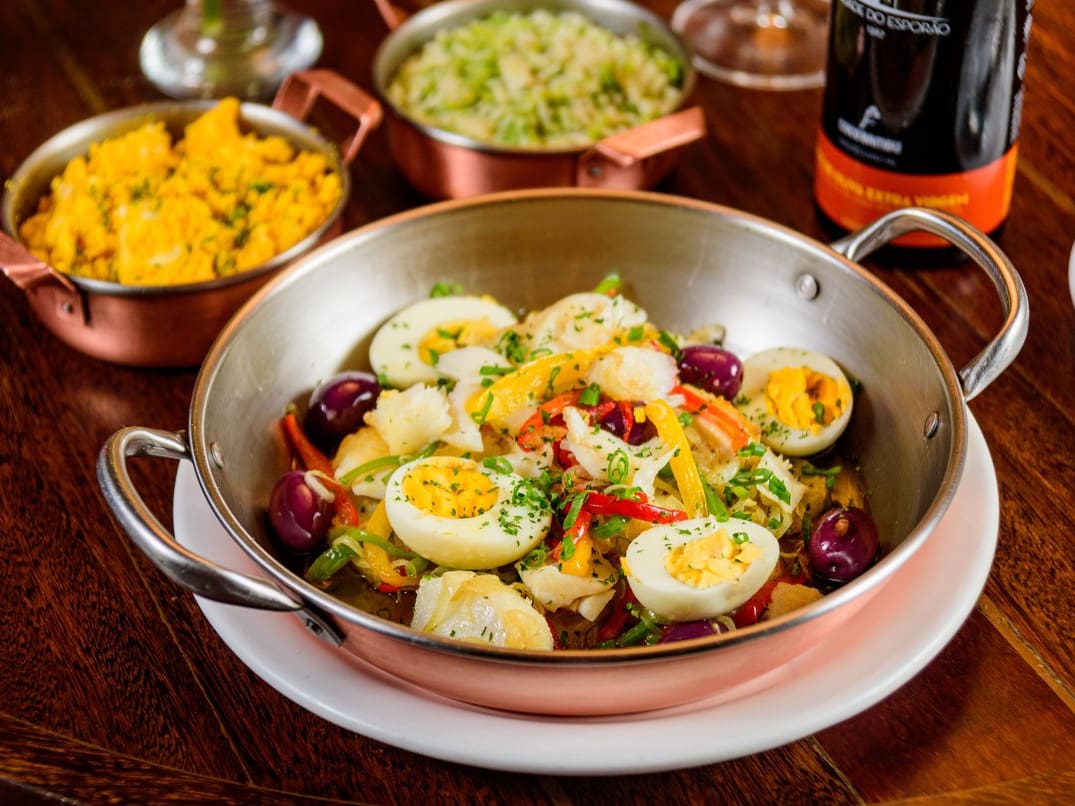Lombo de bacalhau em lascas assado com cebola, mix de pimentões, batatas, ovos, cebolinha, e azeitonas azapa. Acompanha arroz de brócolis e farofa de ovos.