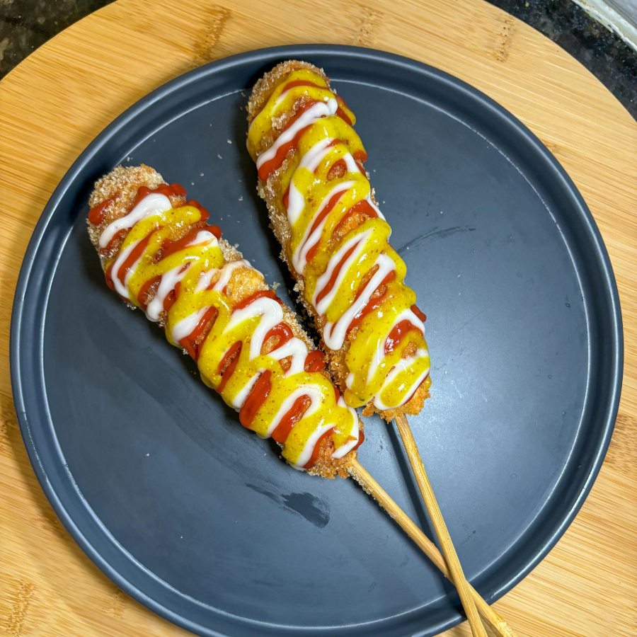 Salsicha empanada com farinha Panko granulada com açúcar e molho de ketchup, maionese e mostarda por cima.