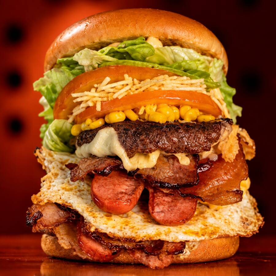 Pão com 2 hambúrgueres(100g), 2 salsichas, 2 queijos, 2 presuntos, bacon crocante, 2 ovos, tomate, alface, milho e batata palha. Um lanche gigante, completo e bem recheado.