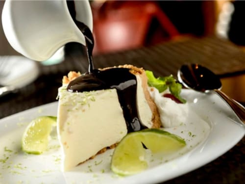 Fatia de torta de creme de limão e leite condensado sobre massa de biscoito. Acompanha calda de chocolate.