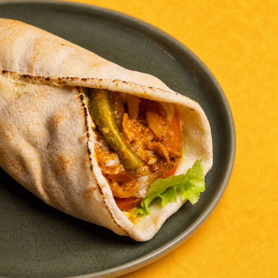 Kebab filé de frango marinado na coalhada seca (contém lactose) com especiarias, pão pitta, hommus tahine, salada de alface e tomate com emulsão de algo e pickles de pepino