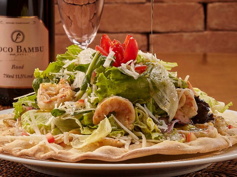 Alface americana, 10 camarões refogados no azeite e ervas, parmesão ralado, molho caesar e croûtons. Servida na casquinha de pizza.