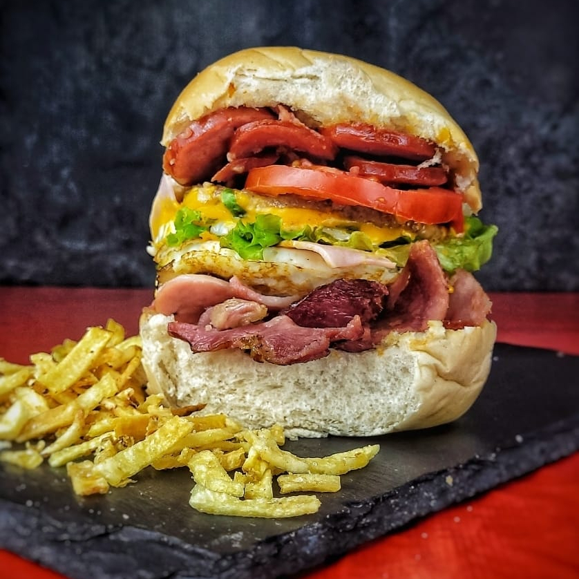 Embarque em uma jornada gastronômica com o nosso X-Duplo, da categoria 'Sublimes Sanduíches'. Este lanche majestoso apresenta um pão bola fresquinho, abrigando duas suculentas camadas de carne, complementadas por dois ovos perfeitamente cozidos. O sabor fica ainda mais intenso com a dupla camada de queijo cheddar derretido e presunto de primeira. Acrescentamos a calabresa crocante e o bacon defumado para uma combinação perfeita. O ketchup e a nossa maionese caseira adicionam um toque especial, enquanto o milho e a ervilha trazem um frescor ao prato. Finalizamos com uma generosa porção de batata palha crocante e uma pitada de queijo ralado. É uma experiência culinária inesquecível!