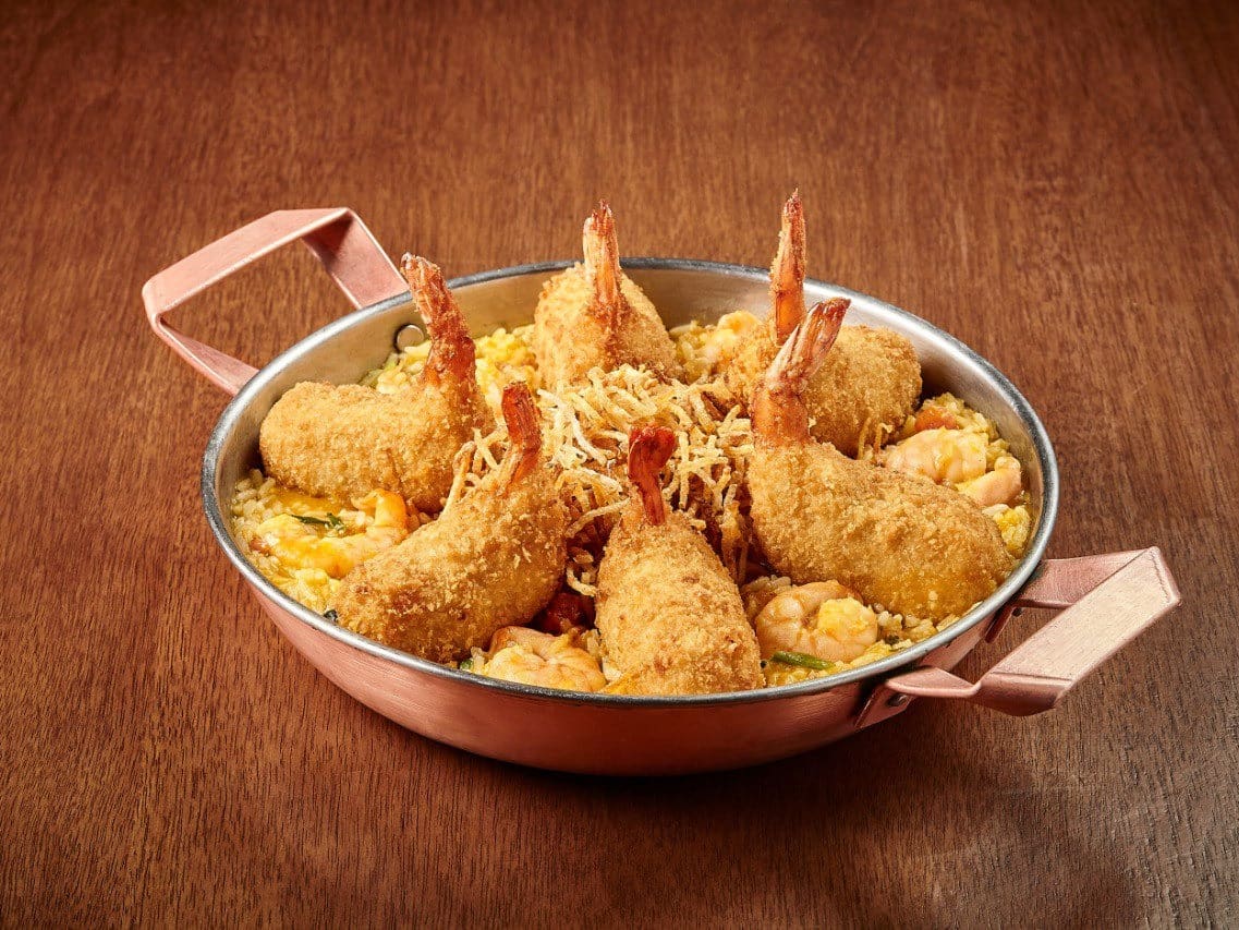 6 Camarões Empanados e Recheados com Catupiry, Sobre Um Cremoso Arroz de Moqueca com Camarões e Coentro, Coberto com Batata Palha. Acompanha Farofa de Panko com Bacon e Alho Poro.