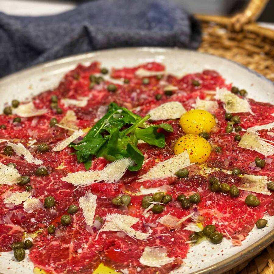 Cama de rúcula, finas fatias de carne bovina (Crua), Mostarda Djon, Alcaparras e Parmesão