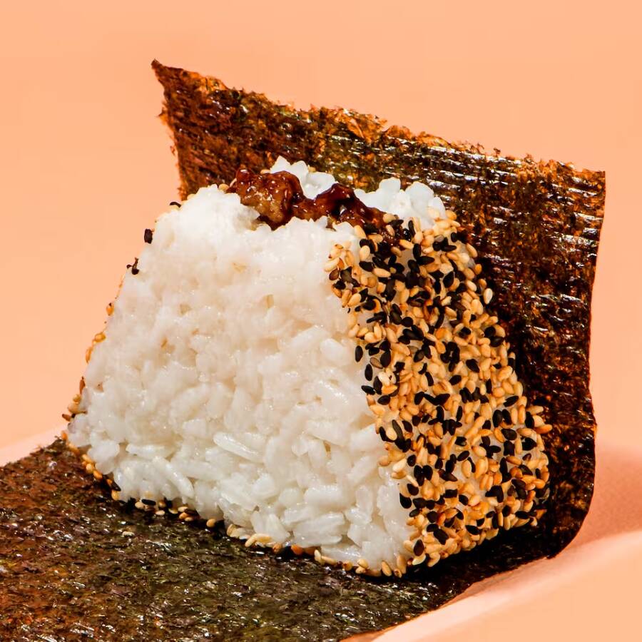 Bolinho de arroz Shari, recheado com berinjela empanada ao molho tonkatsu, envolto por um mix de gergelim e acompanhado de alga nori, embalada separadamente para manter a crocância até o momento de consumo.