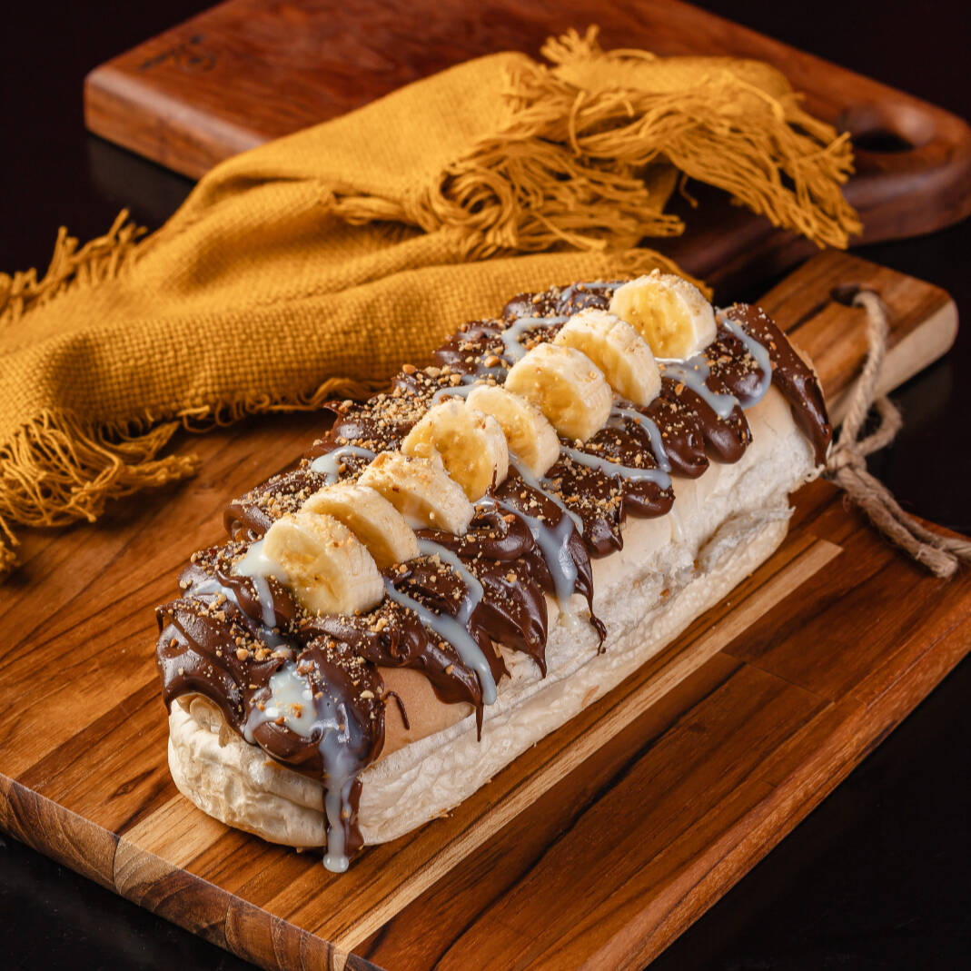 Pão macio de Hot Dog, leite condensado, Banana, canela, farofa de amendoim, Bolinhas de chocolate crocante e chocolate preto derretido.